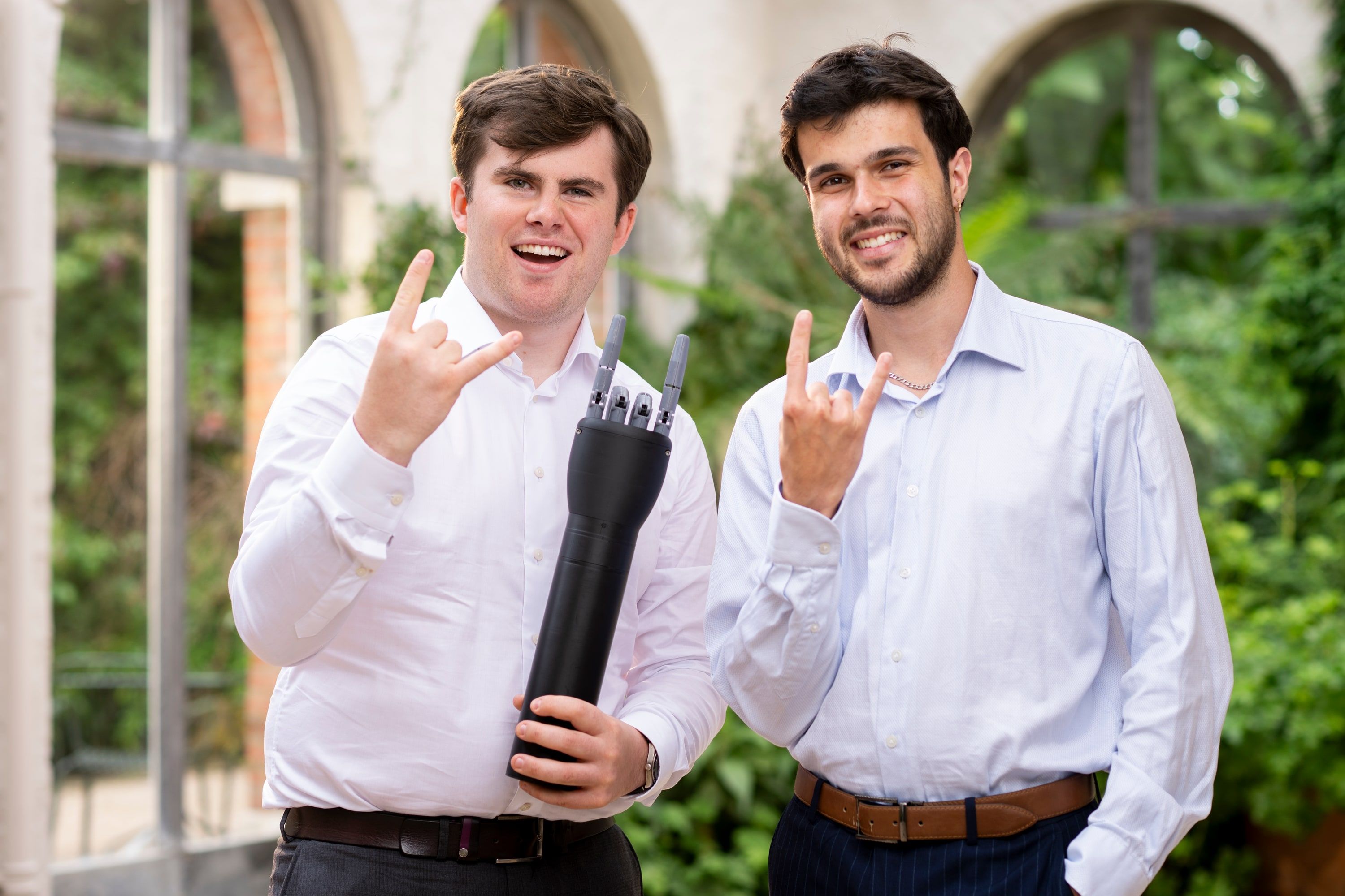 Seán Barrett and William Hartley, smiling and making a rock-on sign, one of them holding Diego, a black prosthetic hand.